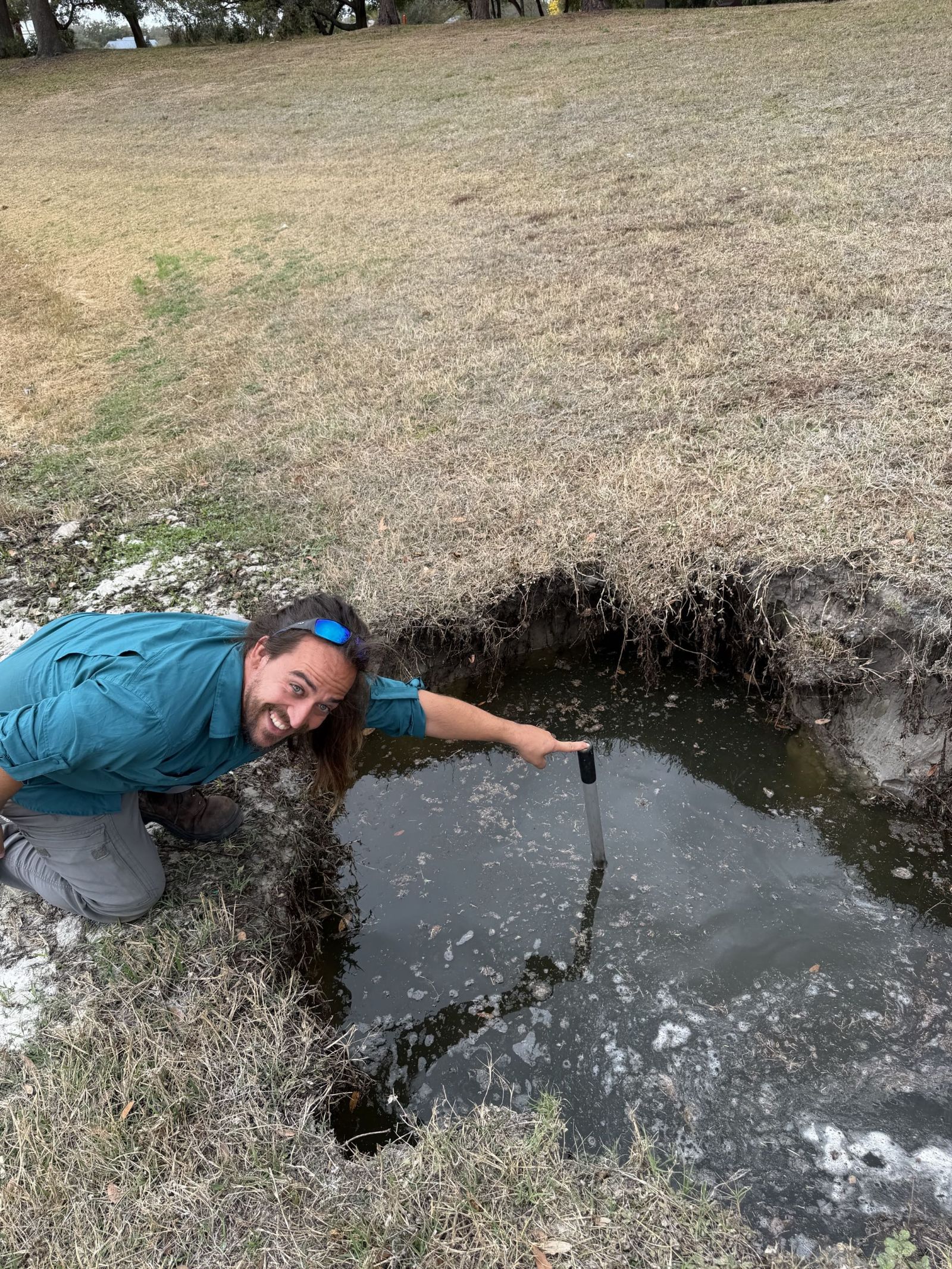 Phillip kneeling beside a flooded hole, smiling and pointing at the broken pipe he just found