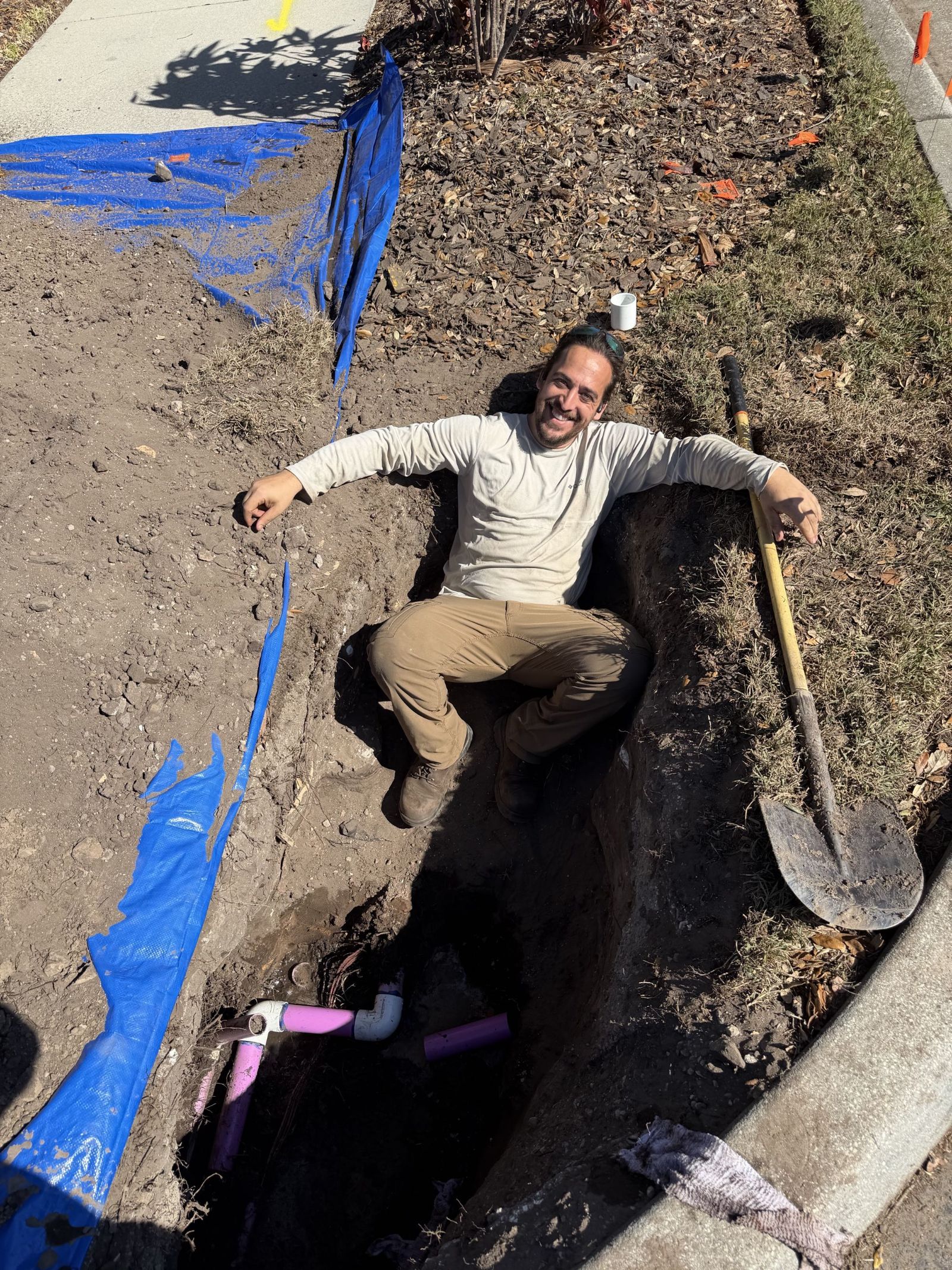 Phillip sitting in a freshly dug trench beside new irrigation piping, smiling at the camera